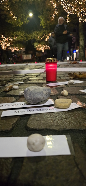 Nahaufnahme von Kopfsteinpflaster bei Nacht, auf dem kleine Steine als Beschwerer auf weißen Papierstreifen mit Namen wie Moritz Mayer liegen. Im Hintergrund leuchtet eine rote Grabkerze, während im unscharfen Hintergrund Menschen unter beleuchteten Bäumen stehen.